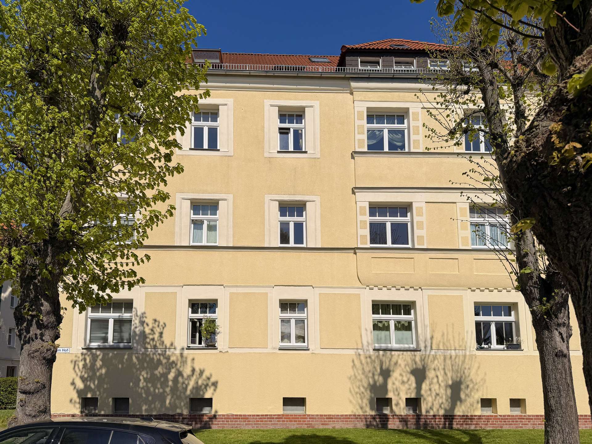 Facade of a pale yellow multi-story building with white-framed windows, flanked by green trees and a blue sky above.