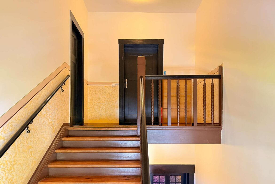 Interior stairwell with wooden steps and dark doorway at the top landing, warm overhead light, and decorative railing on the right side.