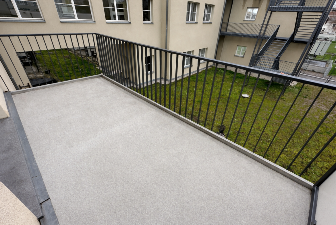 Concrete balcony with a black metal railing, overlooking a grassy courtyard and beige apartment building with an external stairwell