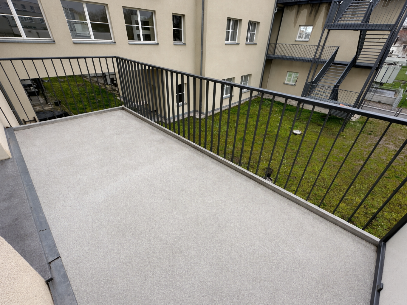Concrete balcony with a black metal railing, overlooking a grassy courtyard and beige apartment building with an external stairwell