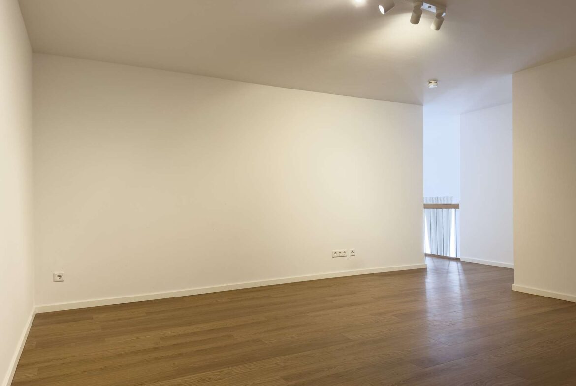 Empty, minimalist interior: white walls, light wood floor, and ceiling track lighting in a spacious room with a doorway to a brighter adjacent area.