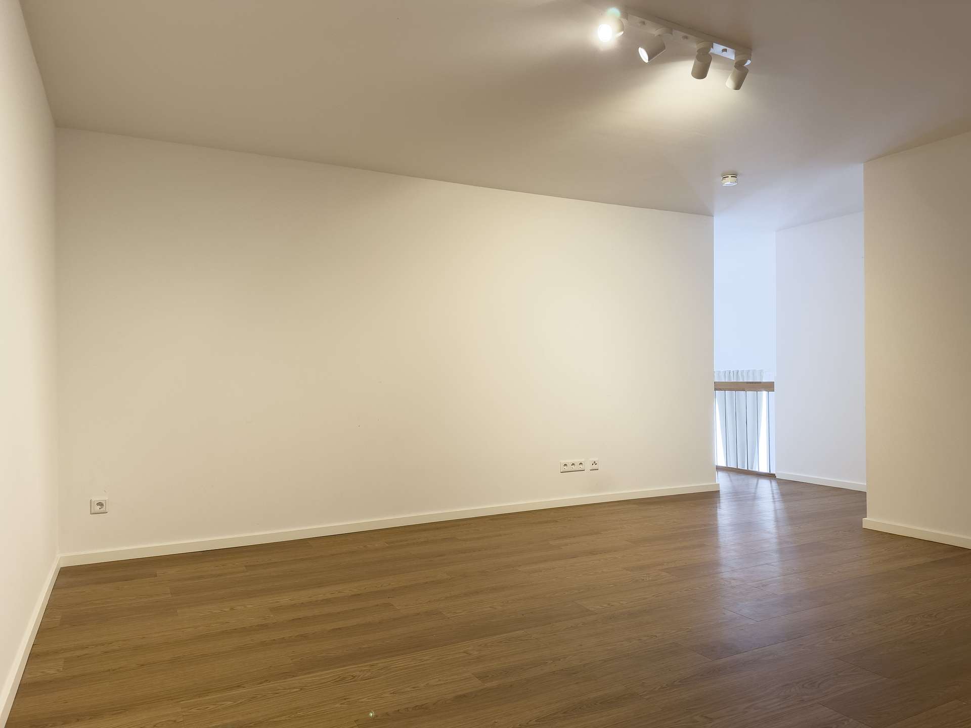 Empty, minimalist interior: white walls, light wood floor, and ceiling track lighting in a spacious room with a doorway to a brighter adjacent area.