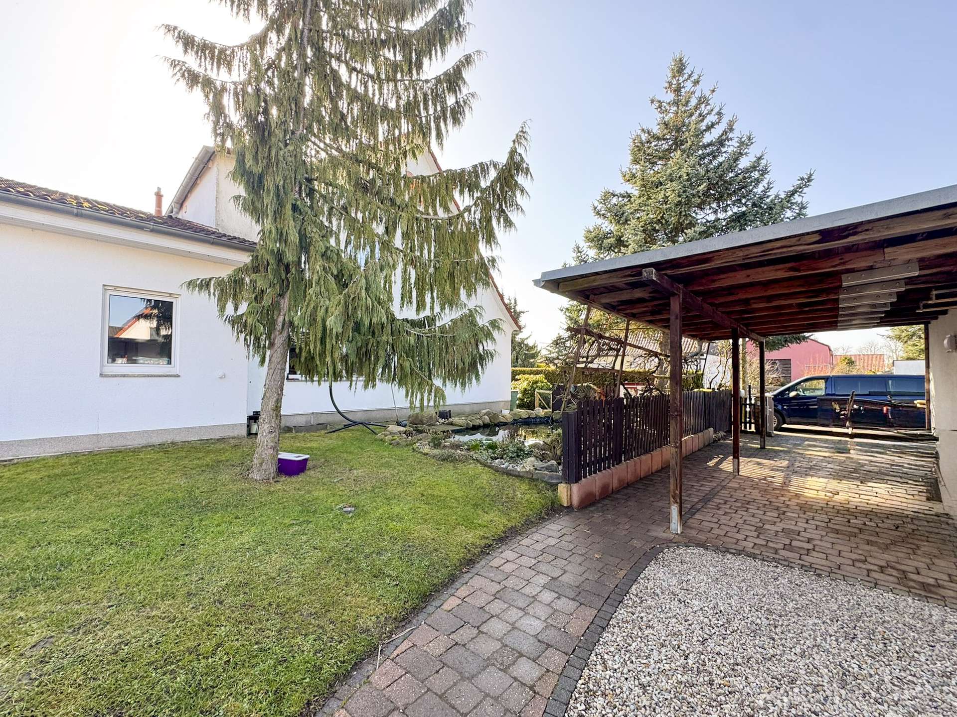 White house with a large evergreen tree in the front yard, window visible on the wall, and a wooden carport on the right with a parked blue van nearby