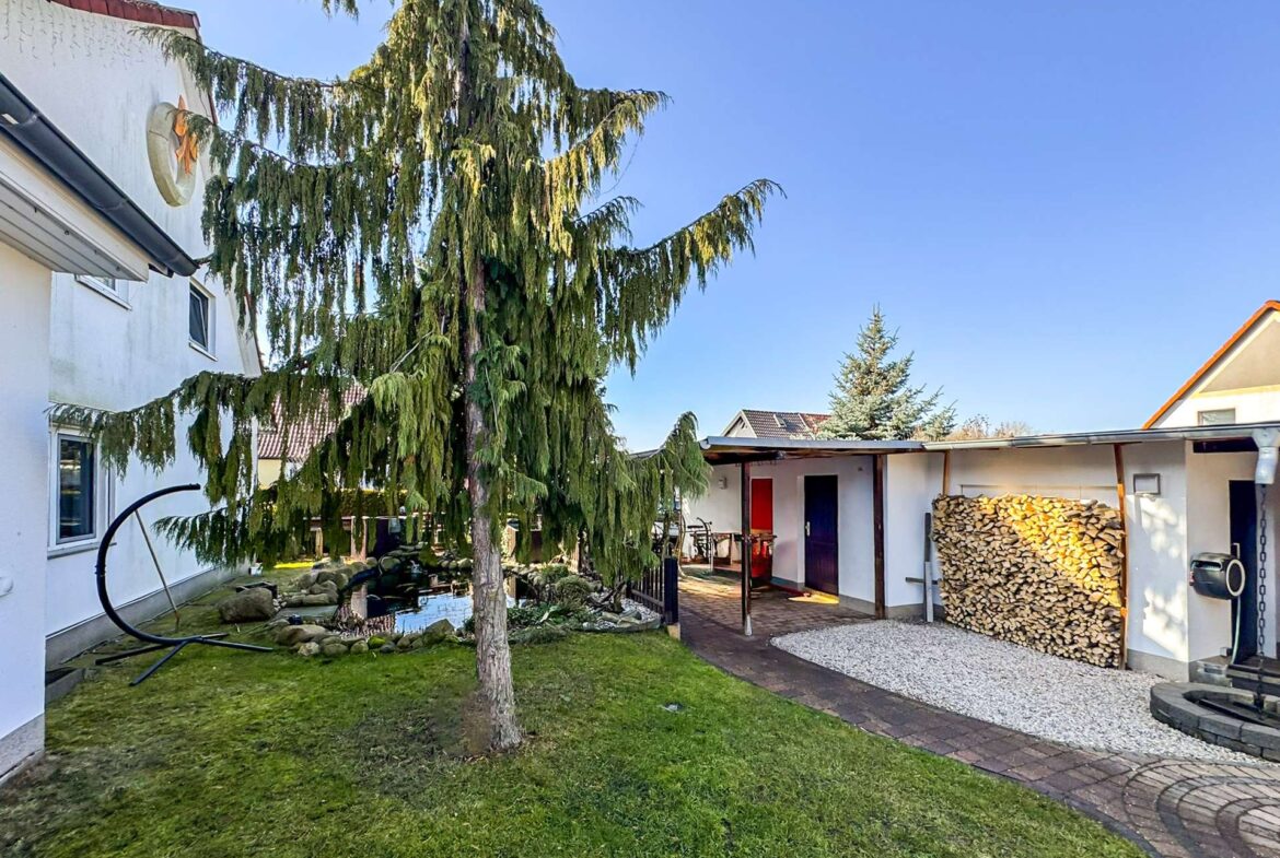 Backyard with a tall evergreen tree near a white house, a covered patio, and a stack of firewood nearby.