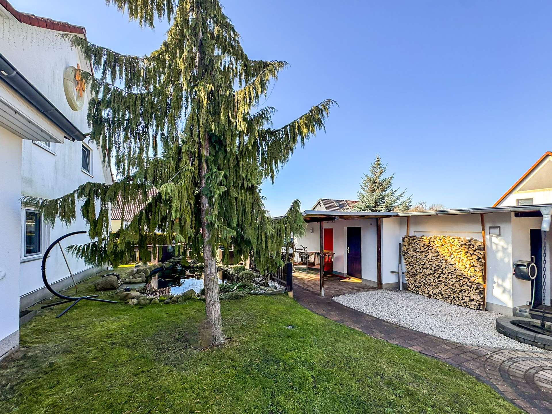 Backyard with a tall evergreen tree near a white house, a covered patio, and a stack of firewood nearby.