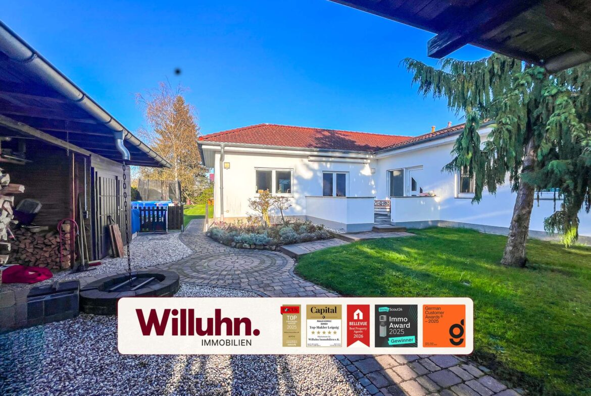 Residential backyard with a white single-story house, red-tiled roof, and a curved stone path; a large Willuhn Immobilien sign with award decals is in the foreground.