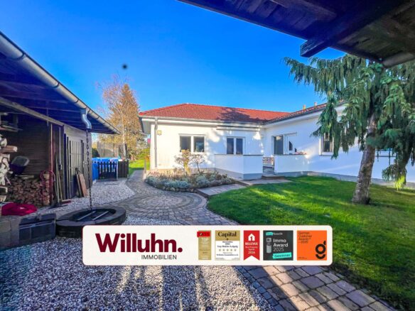 Residential backyard with a white single-story house, red-tiled roof, and a curved stone path; a large Willuhn Immobilien sign with award decals is in the foreground.