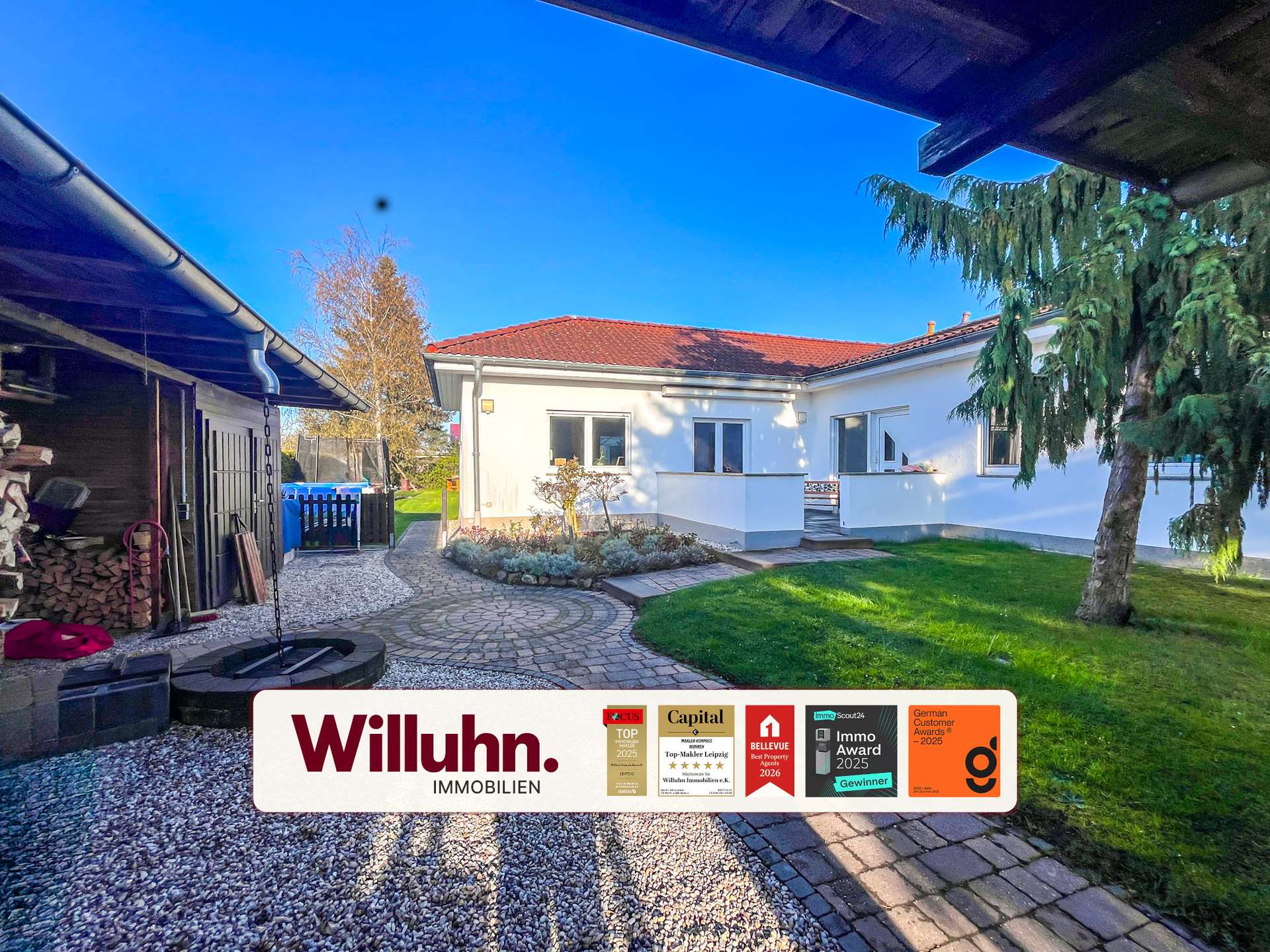 Residential backyard with a white single-story house, red-tiled roof, and a curved stone path; a large Willuhn Immobilien sign with award decals is in the foreground.