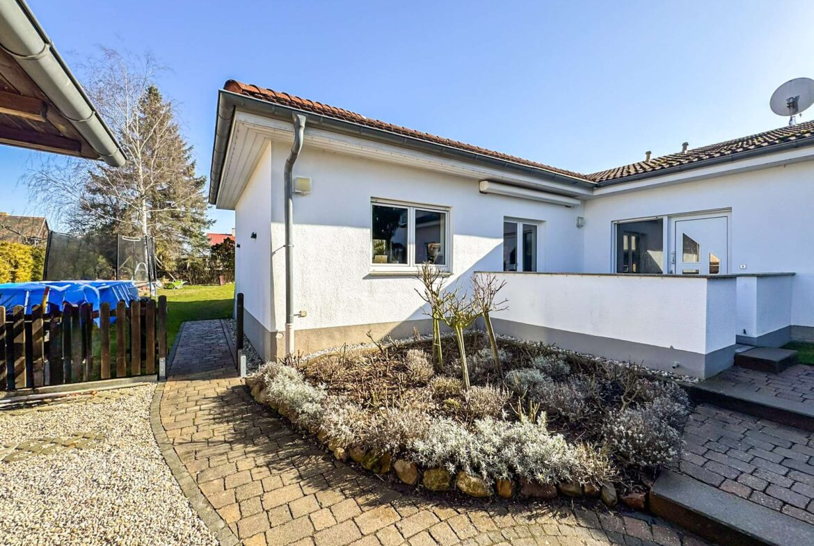 White single-story house with a red-tiled roof, glass door and window, and a paved courtyard curving around a stone planter bed with shrubs.