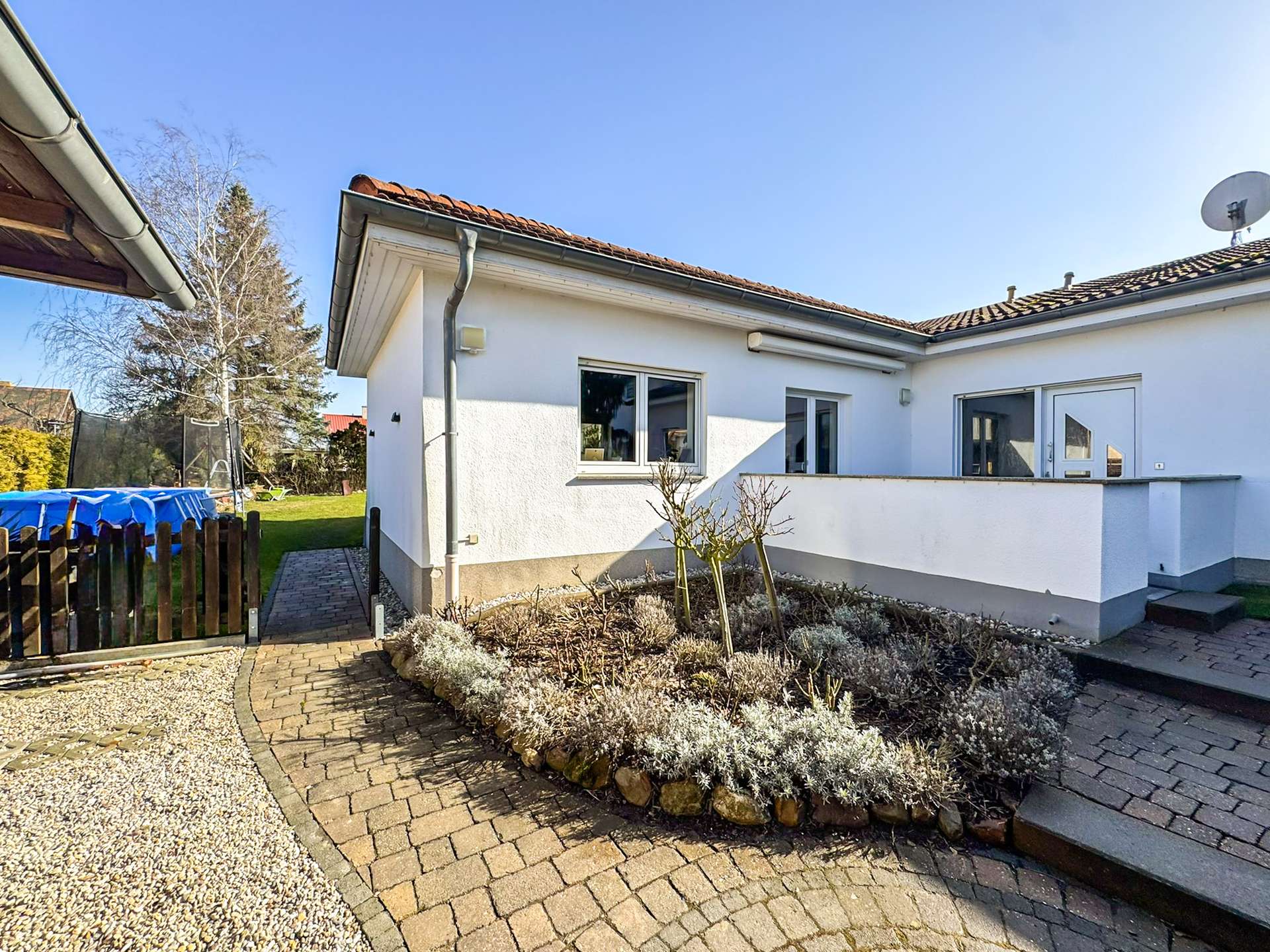 White single-story house with a red-tiled roof, glass door and window, and a paved courtyard curving around a stone planter bed with shrubs.