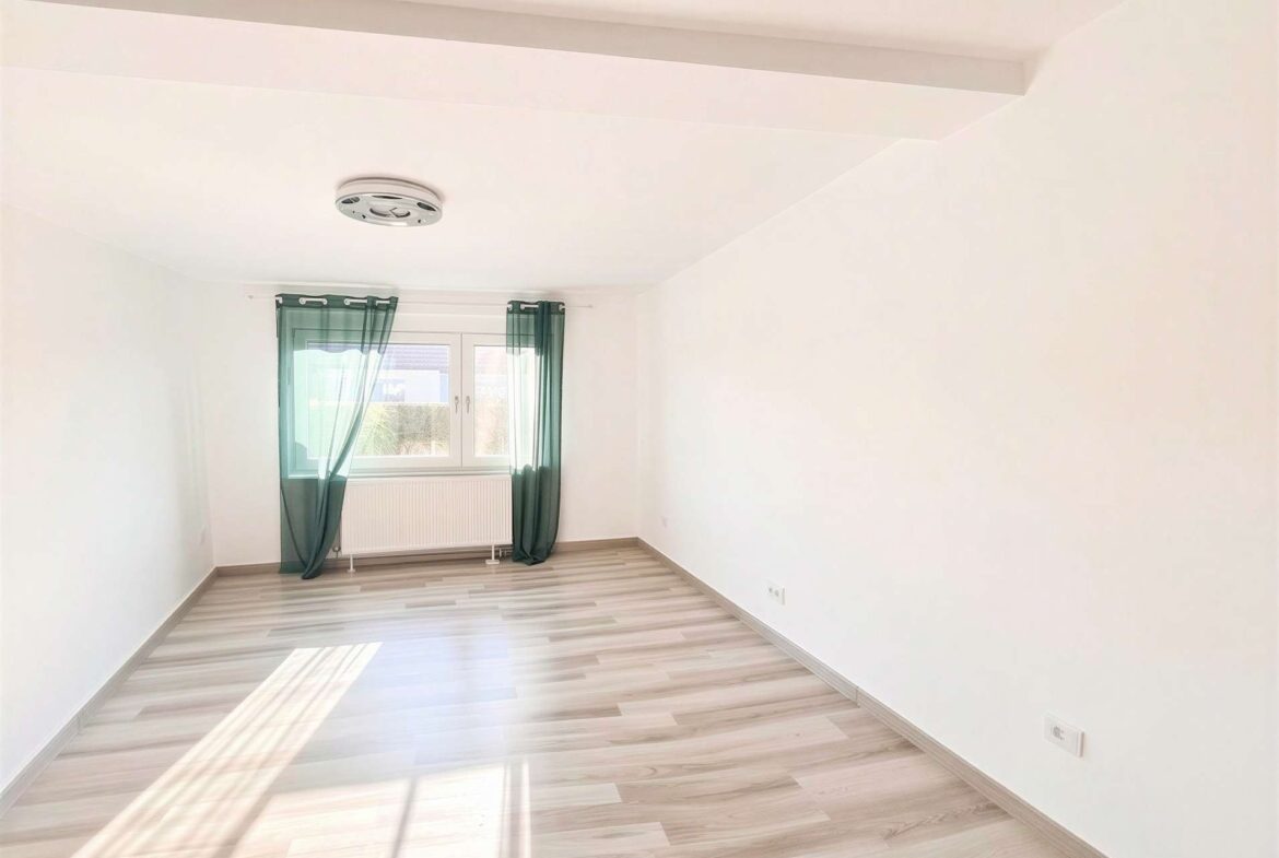 Bright empty room with light wood flooring, white walls, and a window with green sheer curtains letting sunlight in.