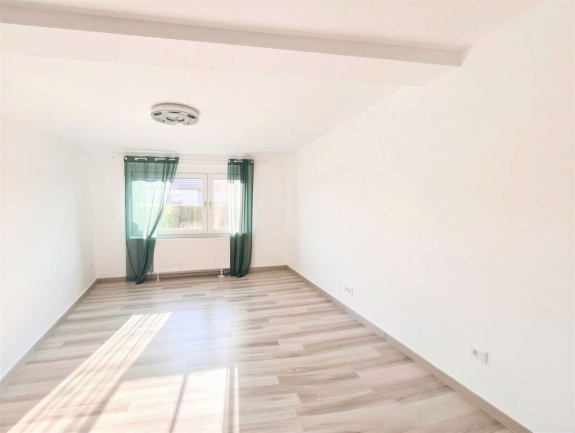 Bright empty room with light wood flooring, white walls, and a window with green sheer curtains letting sunlight in.