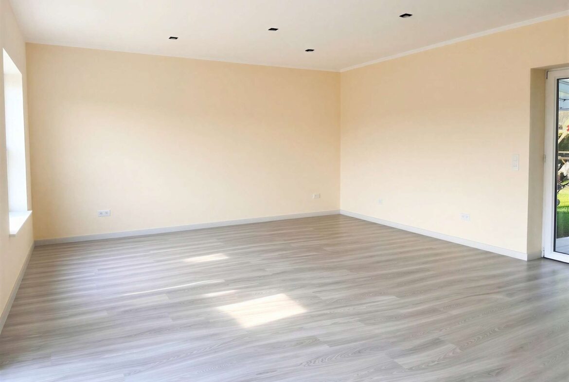 Empty room with pale beige walls, light wood-look flooring, and a row of inset square ceiling lights; a glass door to the outside on the right joins a windowed wall on the left.