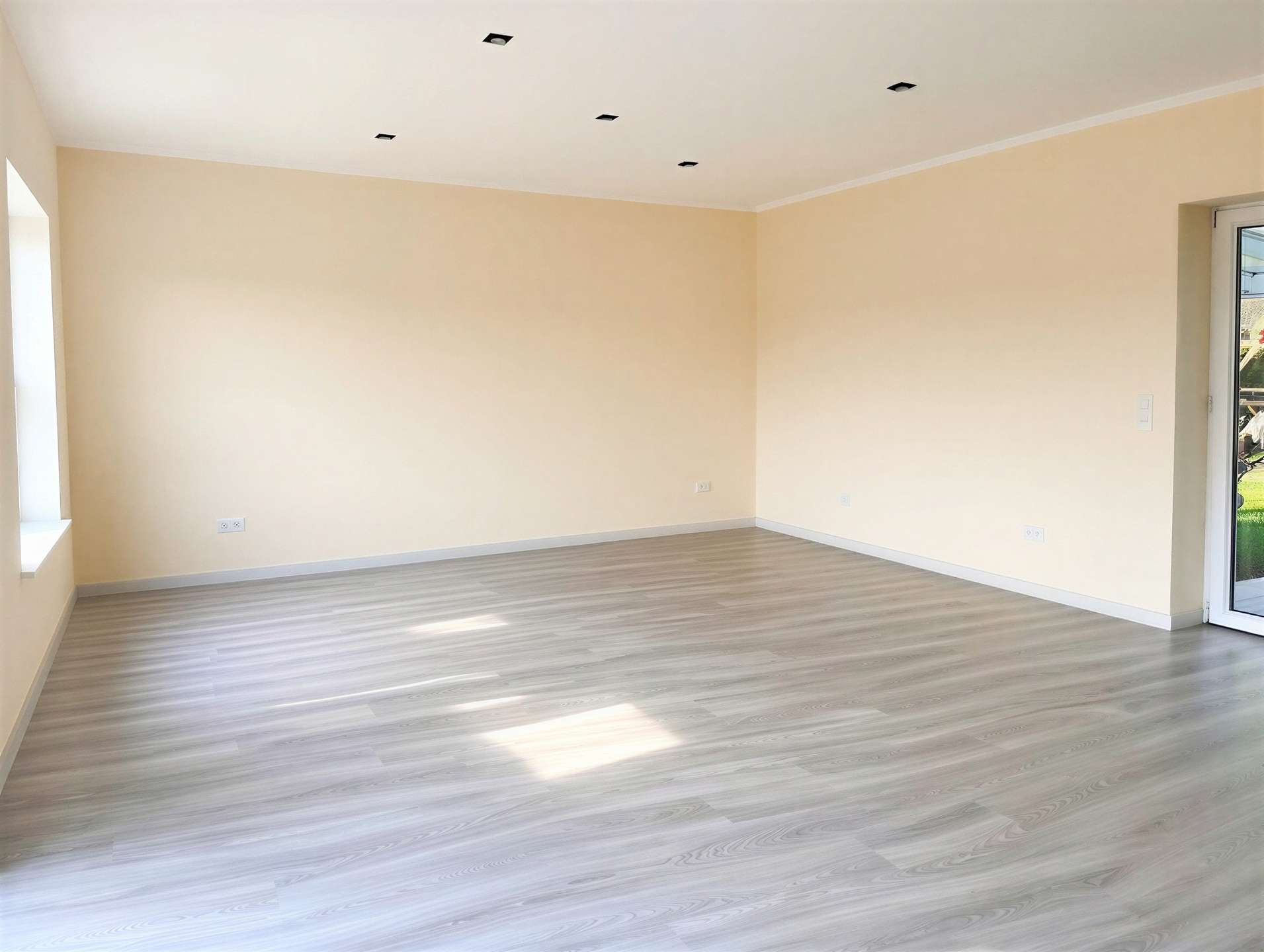 Empty room with pale beige walls, light wood-look flooring, and a row of inset square ceiling lights; a glass door to the outside on the right joins a windowed wall on the left.