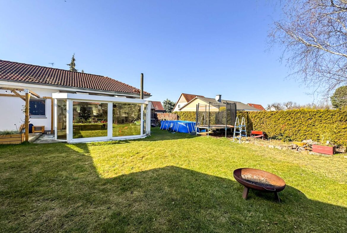 Backyard with a glass-walled sunroom attached to a white house, trampoline, and a blue pool in a fenced yard on a sunny day.