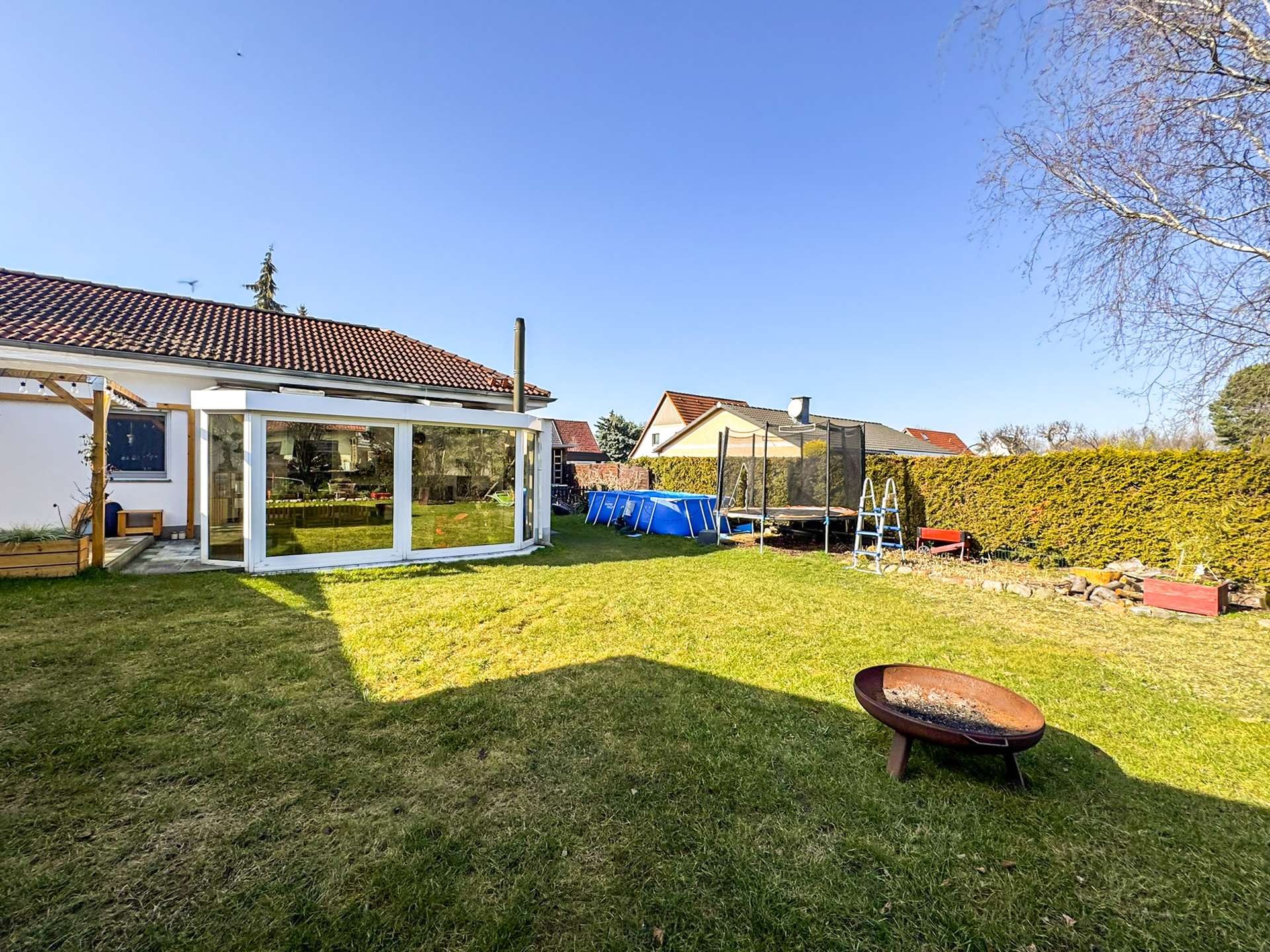 Backyard with a glass-walled sunroom attached to a white house, trampoline, and a blue pool in a fenced yard on a sunny day.