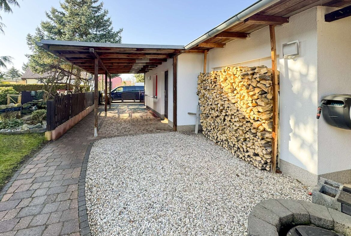 Driveway under a wooden carport beside a white house; large stack of firewood and a parked car in the background.