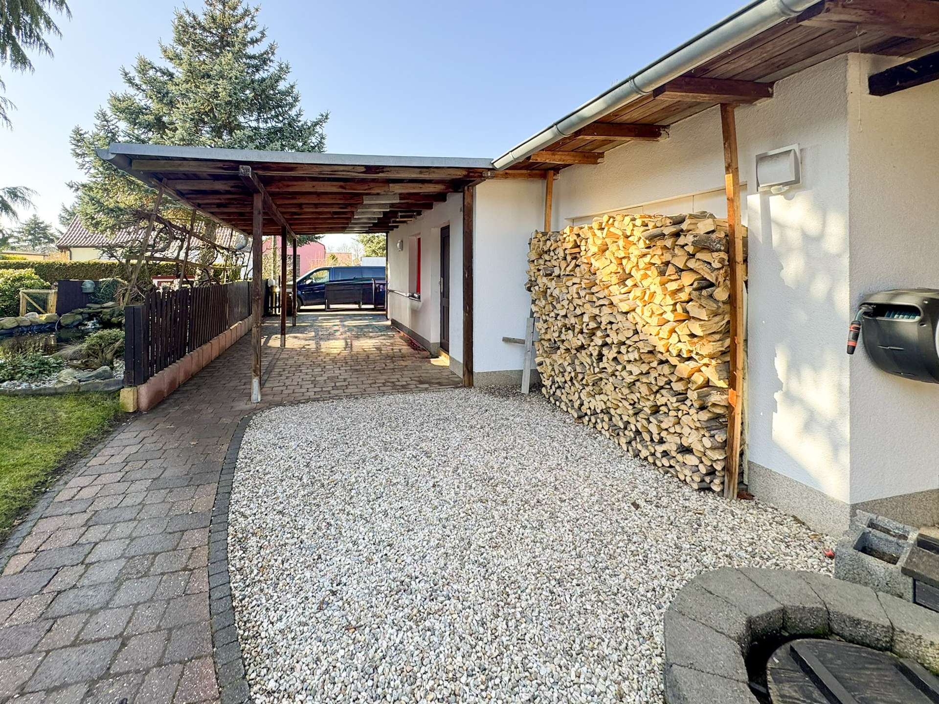 Driveway under a wooden carport beside a white house; large stack of firewood and a parked car in the background.