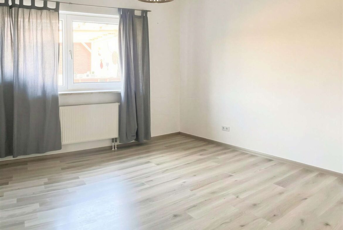 Empty room with a large window covered by gray curtains, radiator beneath the window, and light wood laminate flooring.