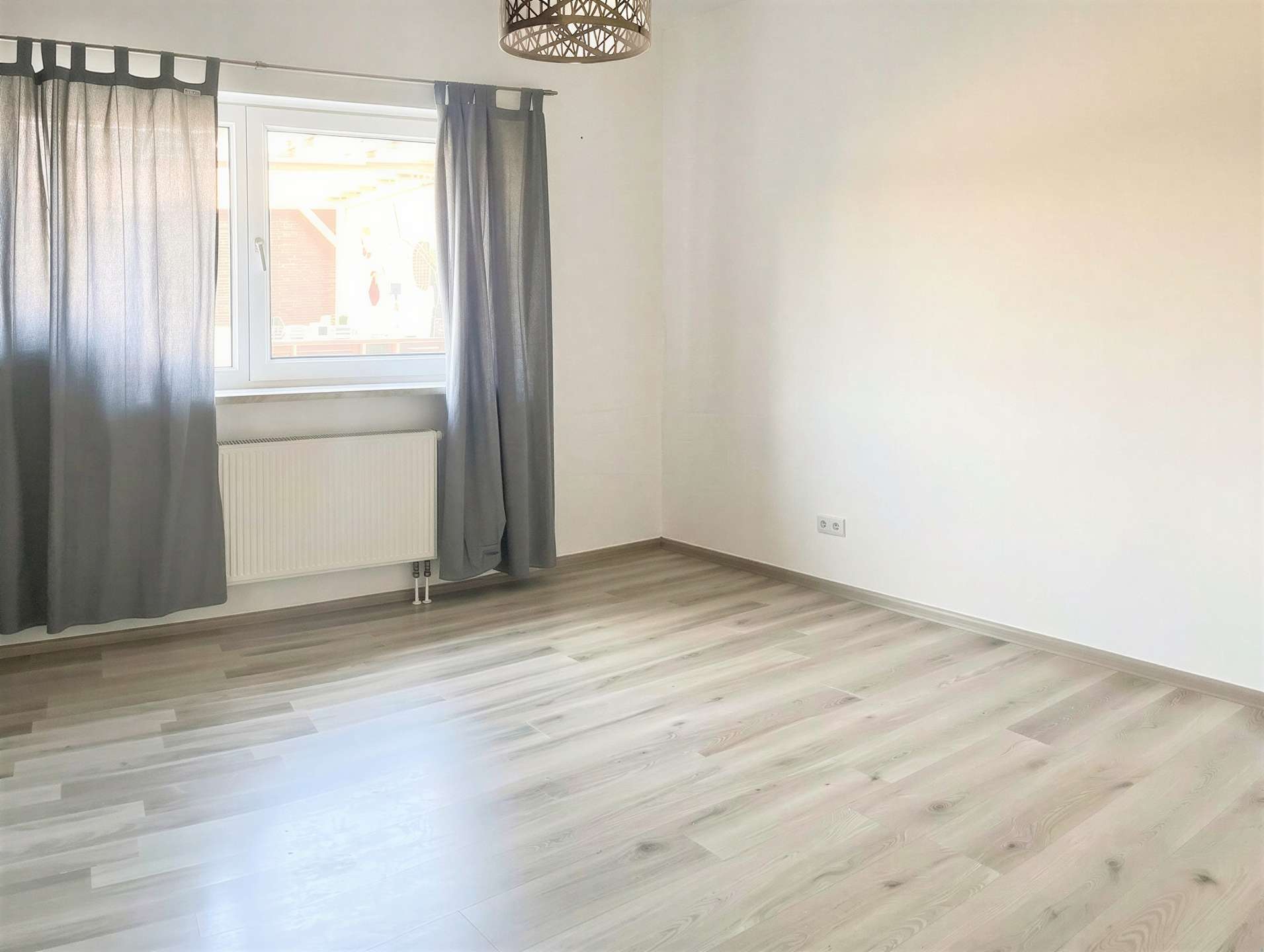 Empty room with a large window covered by gray curtains, radiator beneath the window, and light wood laminate flooring.