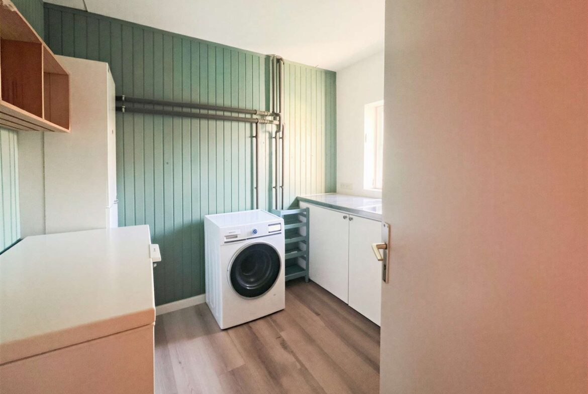 Laundry room with a front-loading washing machine, white countertops, and pale green paneled walls; exposed pipes run along the wall and a window lets in light.