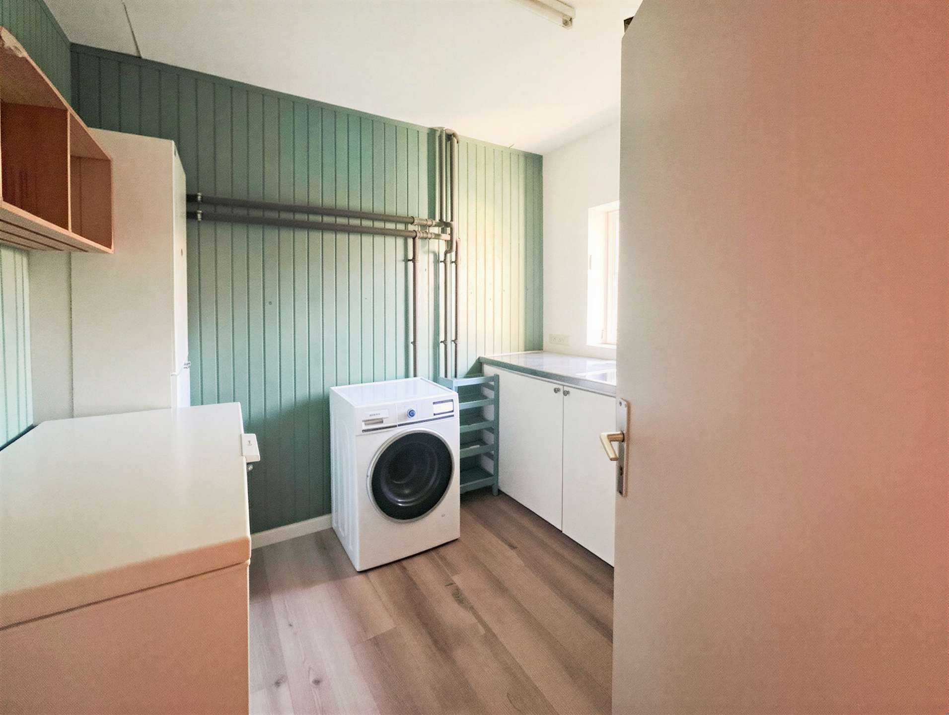 Laundry room with a front-loading washing machine, white countertops, and pale green paneled walls; exposed pipes run along the wall and a window lets in light.