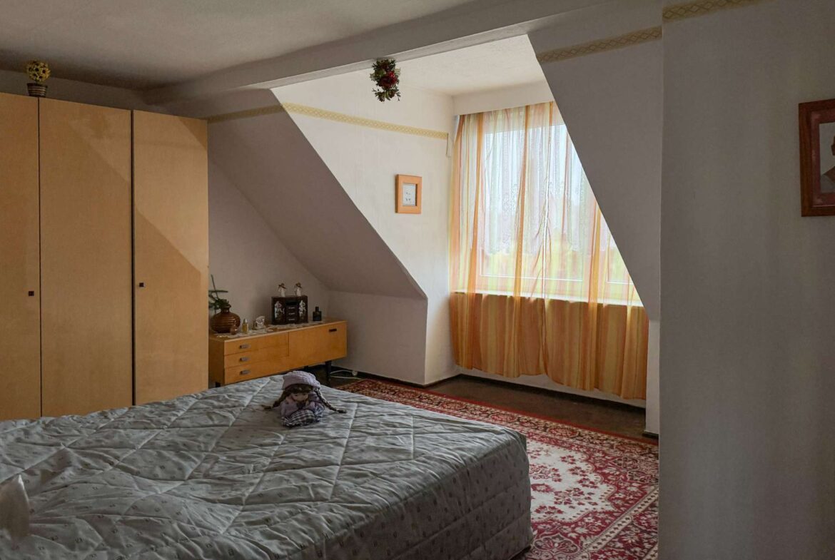 Bedroom with a large bed in the foreground, grey quilt, wooden wardrobes on the left, and a sunlit window with orange curtains in a sloped alcove.
