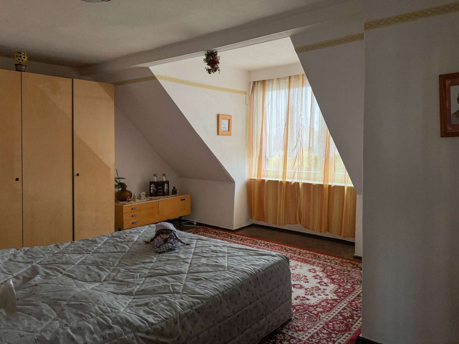 Bedroom with a large bed in the foreground, grey quilt, wooden wardrobes on the left, and a sunlit window with orange curtains in a sloped alcove.