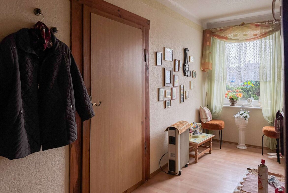 Coat hanging on a wall hook beside a wooden door in a cozy hallway with framed photos and a decorative clock on the adjacent wall, near a window.