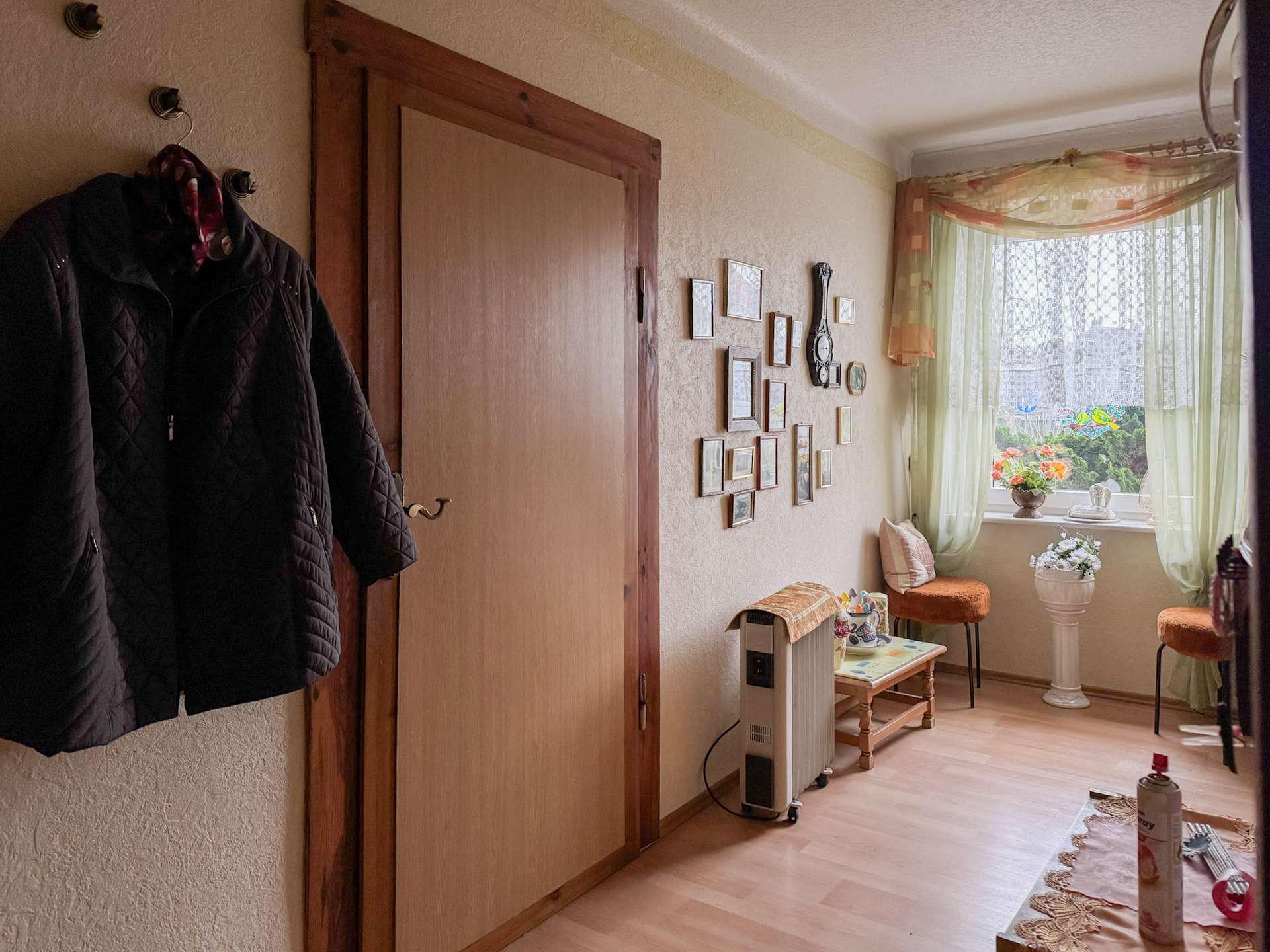Coat hanging on a wall hook beside a wooden door in a cozy hallway with framed photos and a decorative clock on the adjacent wall, near a window.