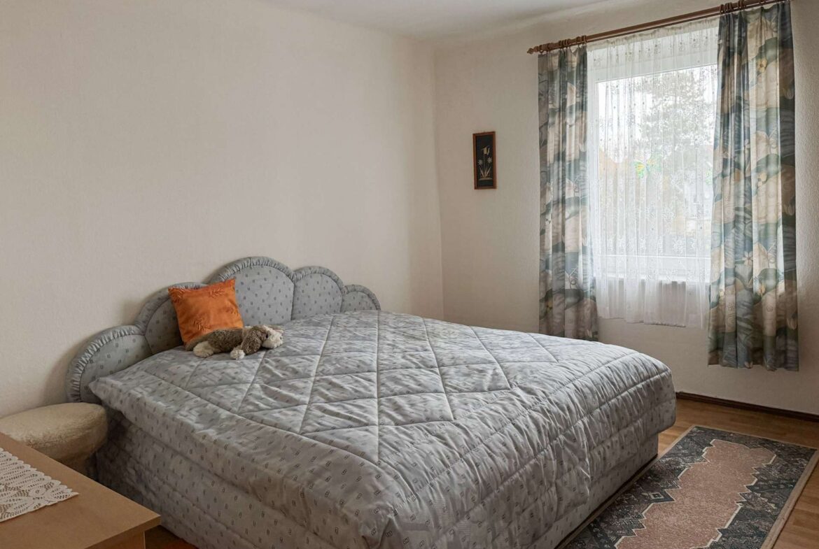 Queen-size bed with a light gray quilt and an orange pillow, beside a nightstand with a toy on it, in a cozy bedroom corner.