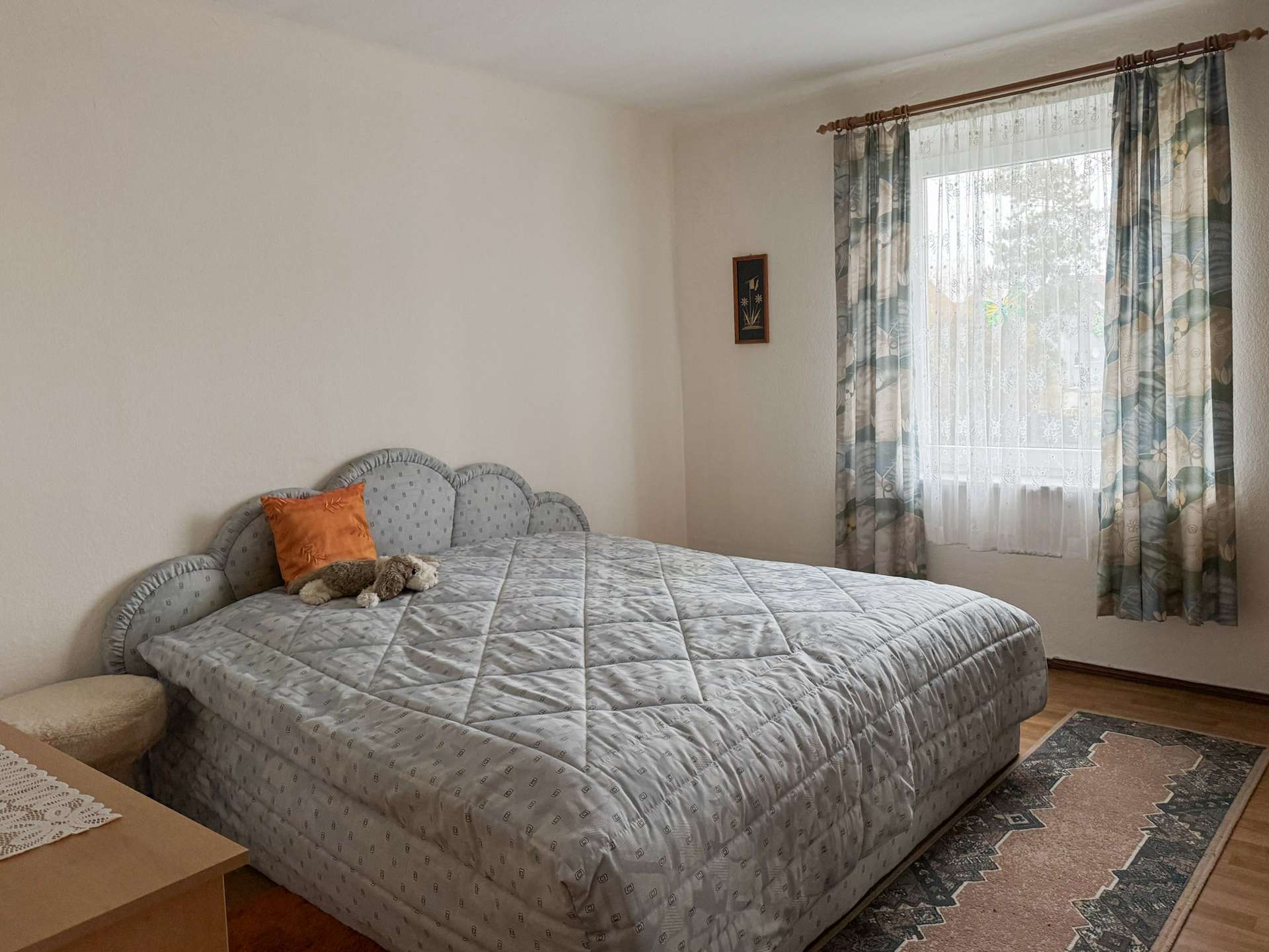 Queen-size bed with a light gray quilt and an orange pillow, beside a nightstand with a toy on it, in a cozy bedroom corner.