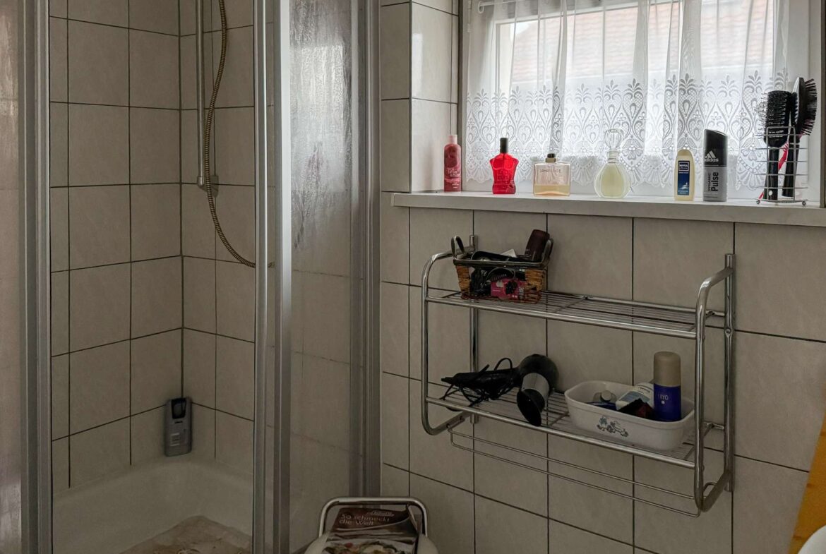 Shower stall on the left with a handheld spray, tiled walls, and a window with a lace curtain above a chrome shelf holding lotions and bottles on the right.