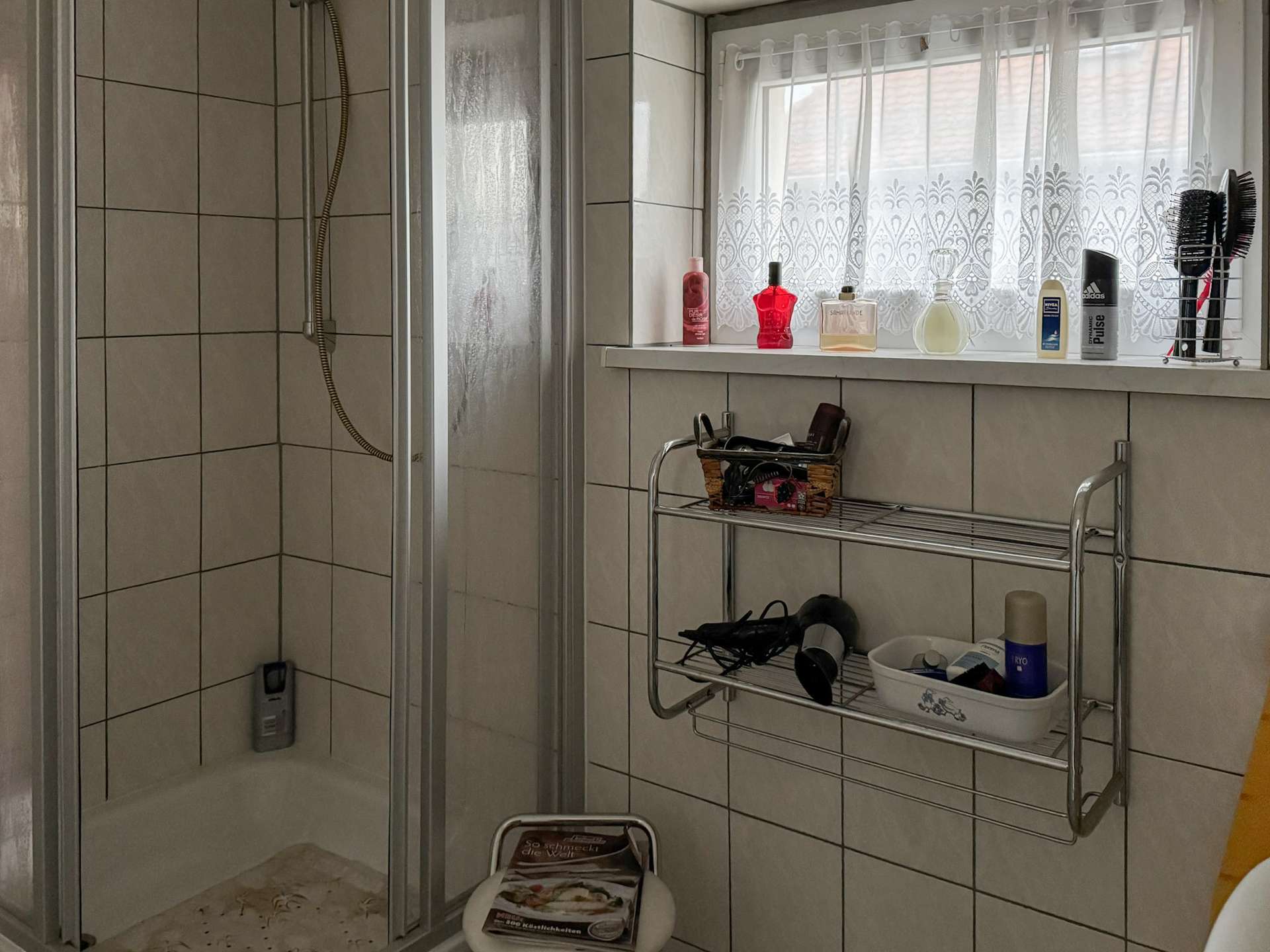 Shower stall on the left with a handheld spray, tiled walls, and a window with a lace curtain above a chrome shelf holding lotions and bottles on the right.