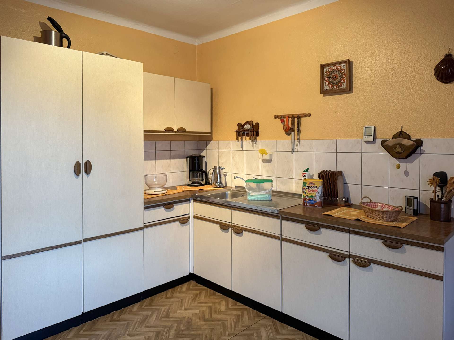 A L-shaped kitchen with white cabinets, beige walls, and tiled backsplash; coffee maker and kettle on the counter.