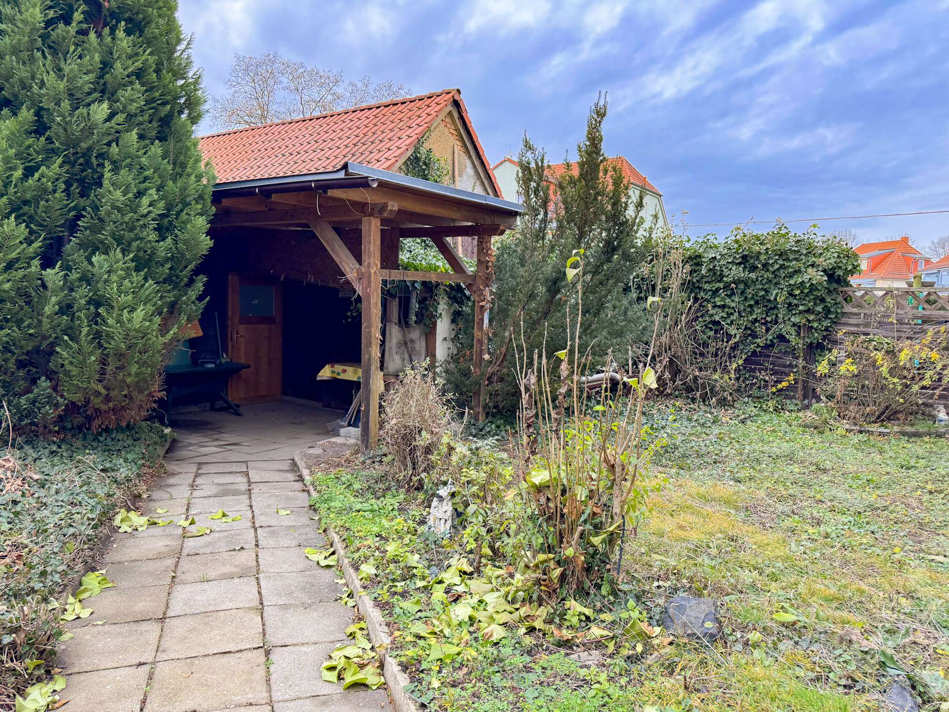 Backyard scene with a wooden shed and tiled roof, surrounded by ivy and overgrown plants along a stone path.