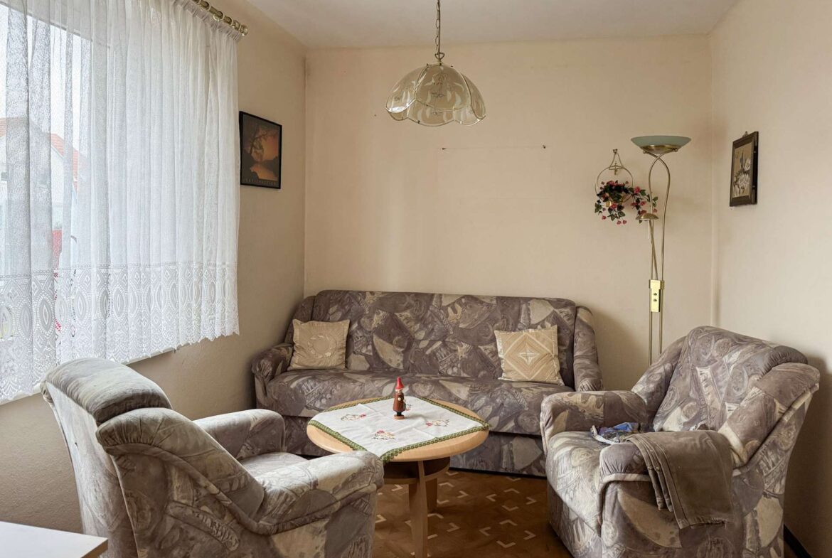 Cozy living room with beige patterned sofa set around a round table, lace curtains on the window, wall art, and a tall floor lamp with a hanging flower basket.