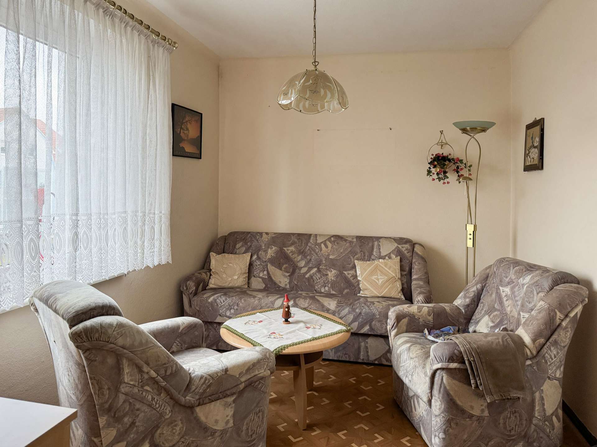 Cozy living room with beige patterned sofa set around a round table, lace curtains on the window, wall art, and a tall floor lamp with a hanging flower basket.