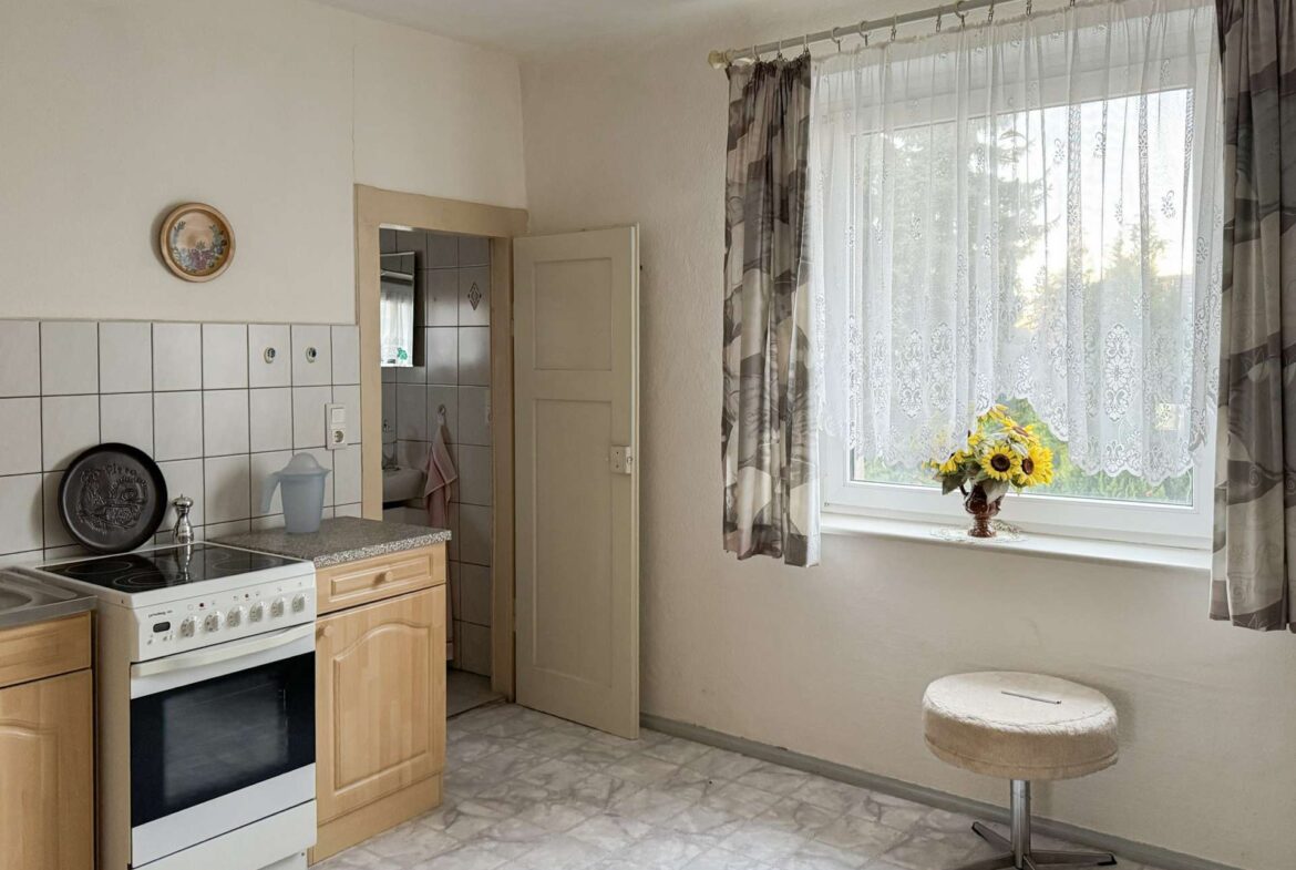 A small kitchen with a white oven, wooden cabinets, and tiled backsplash, next to a doorway leading to another room; a window with lace curtains and sunflowers on the sill nearby.