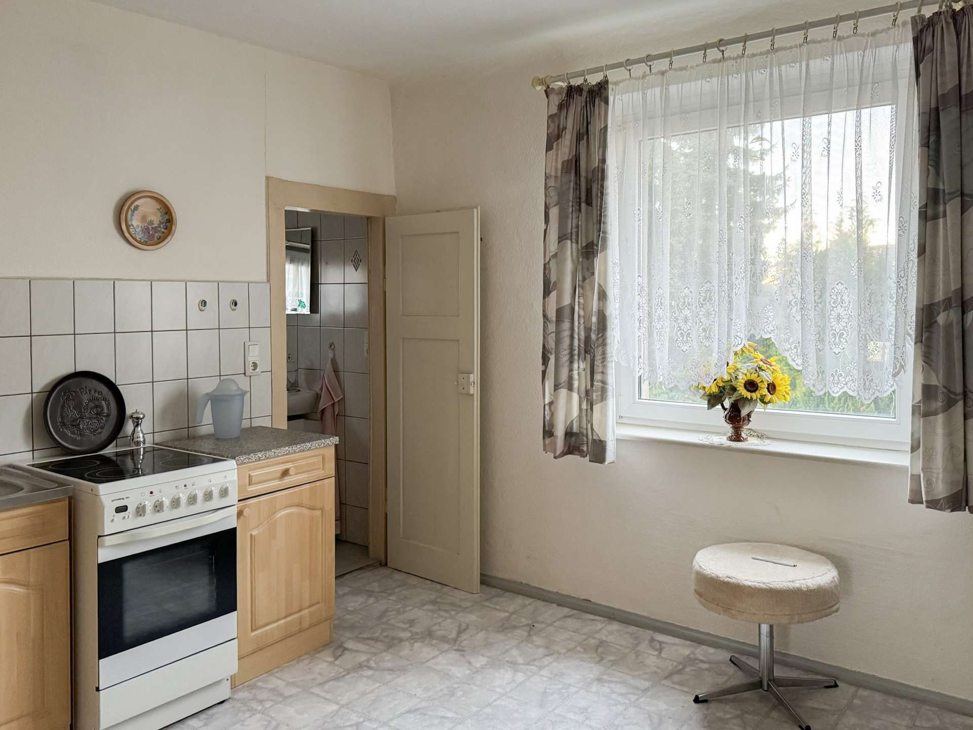 A small kitchen with a white oven, wooden cabinets, and tiled backsplash, next to a doorway leading to another room; a window with lace curtains and sunflowers on the sill nearby.