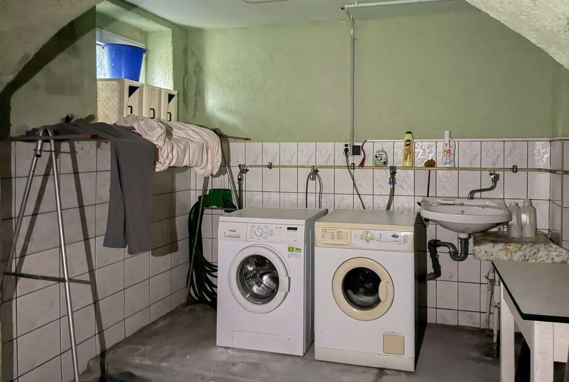 Basement laundry area with a washing machine and dryer side by side, towels and clothes draped over a rack, and cleaning supplies on a tiled counter shelf behind them.