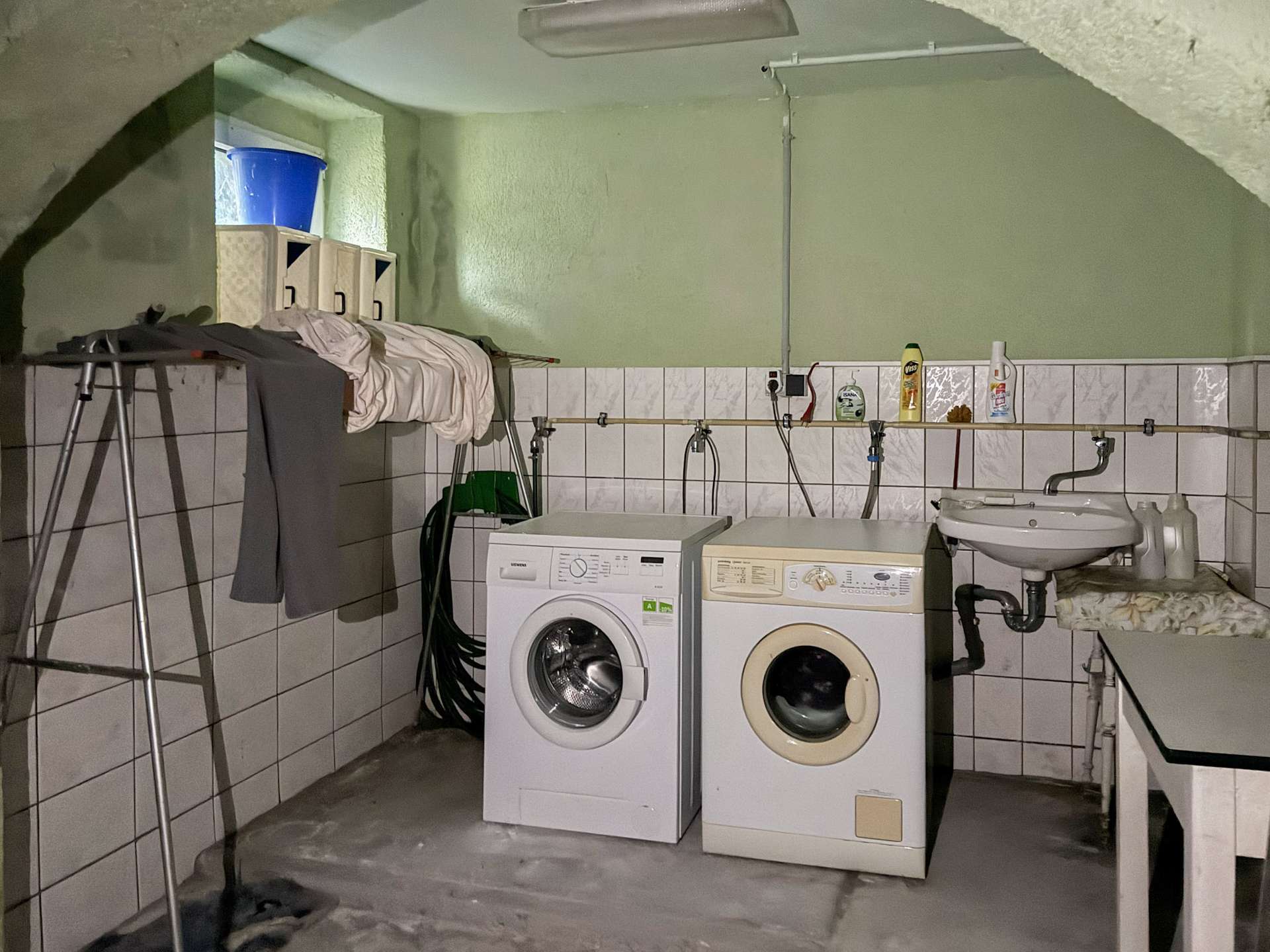 Basement laundry area with a washing machine and dryer side by side, towels and clothes draped over a rack, and cleaning supplies on a tiled counter shelf behind them.