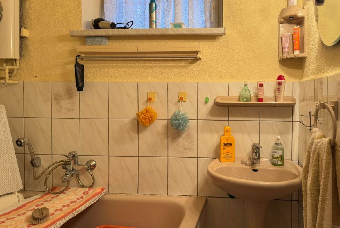 Small bathroom with a white sink and bathtub, yellow walls, and tiled wainscoting; shelves hold shampoo and lotion bottles near a window with lace curtain.