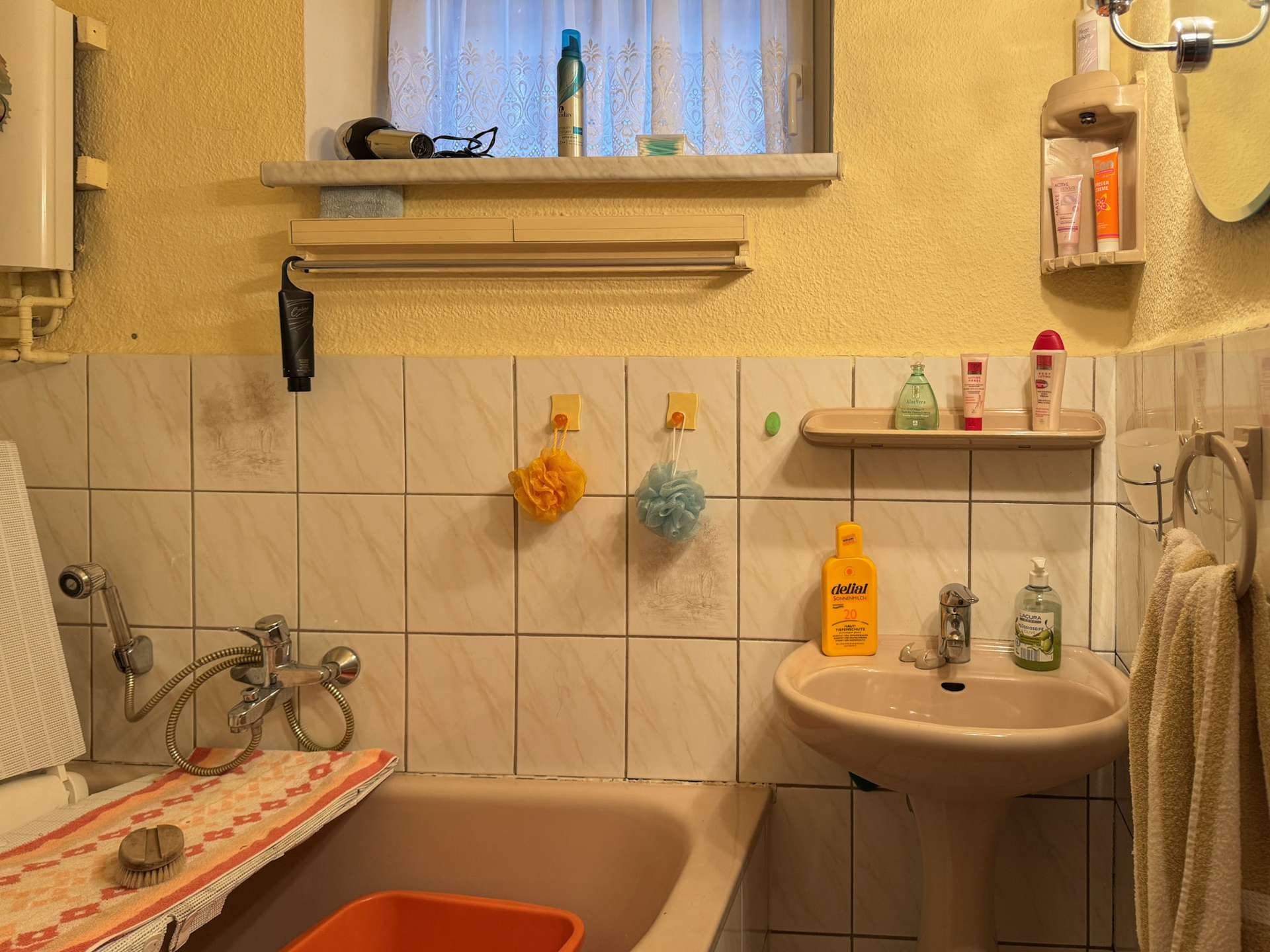 Small bathroom with a white sink and bathtub, yellow walls, and tiled wainscoting; shelves hold shampoo and lotion bottles near a window with lace curtain.