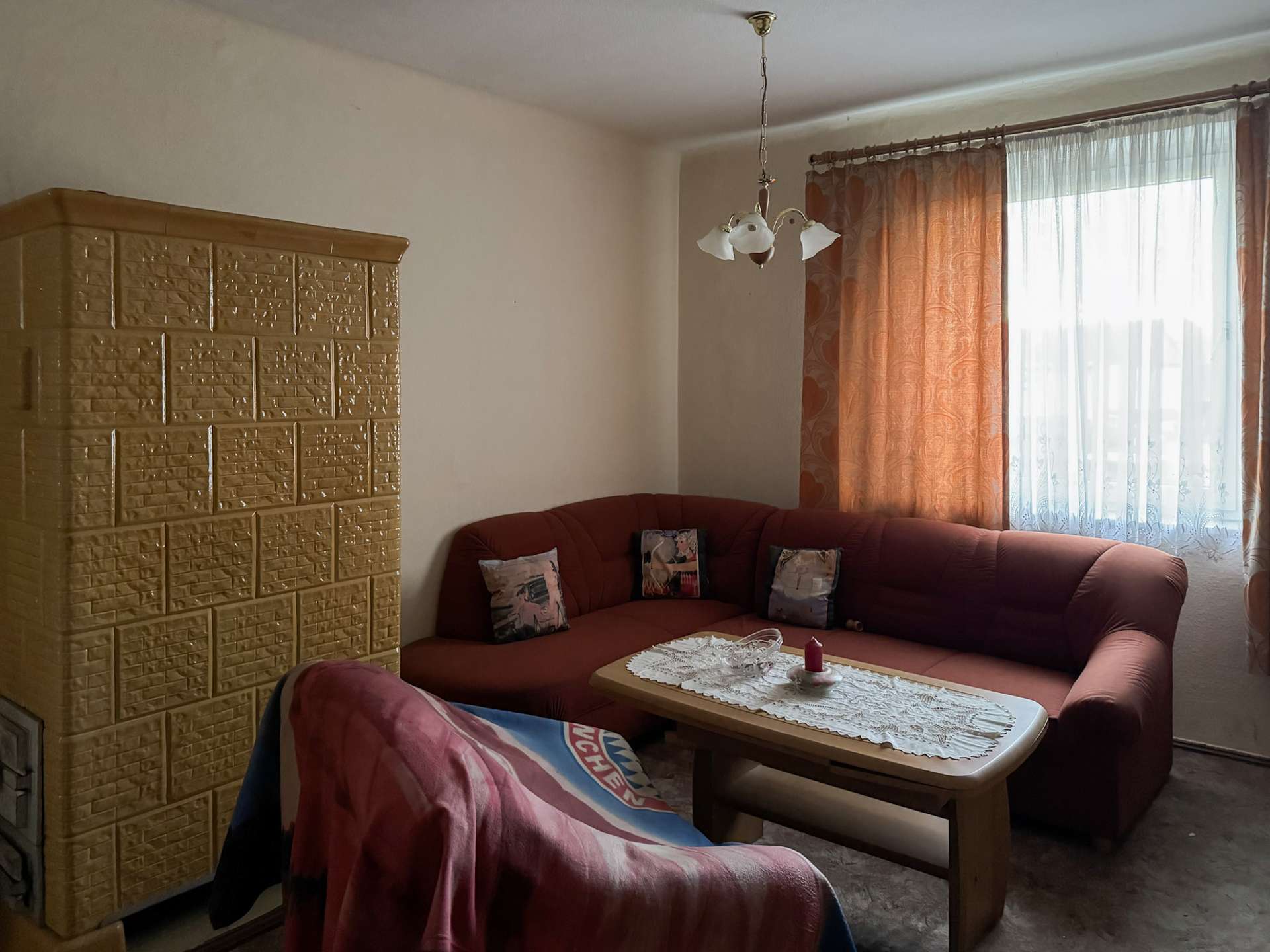 Cozy living room corner with a red sectional sofa, decorative cushions, and a wooden coffee table by a large window with sheer white and orange curtains.