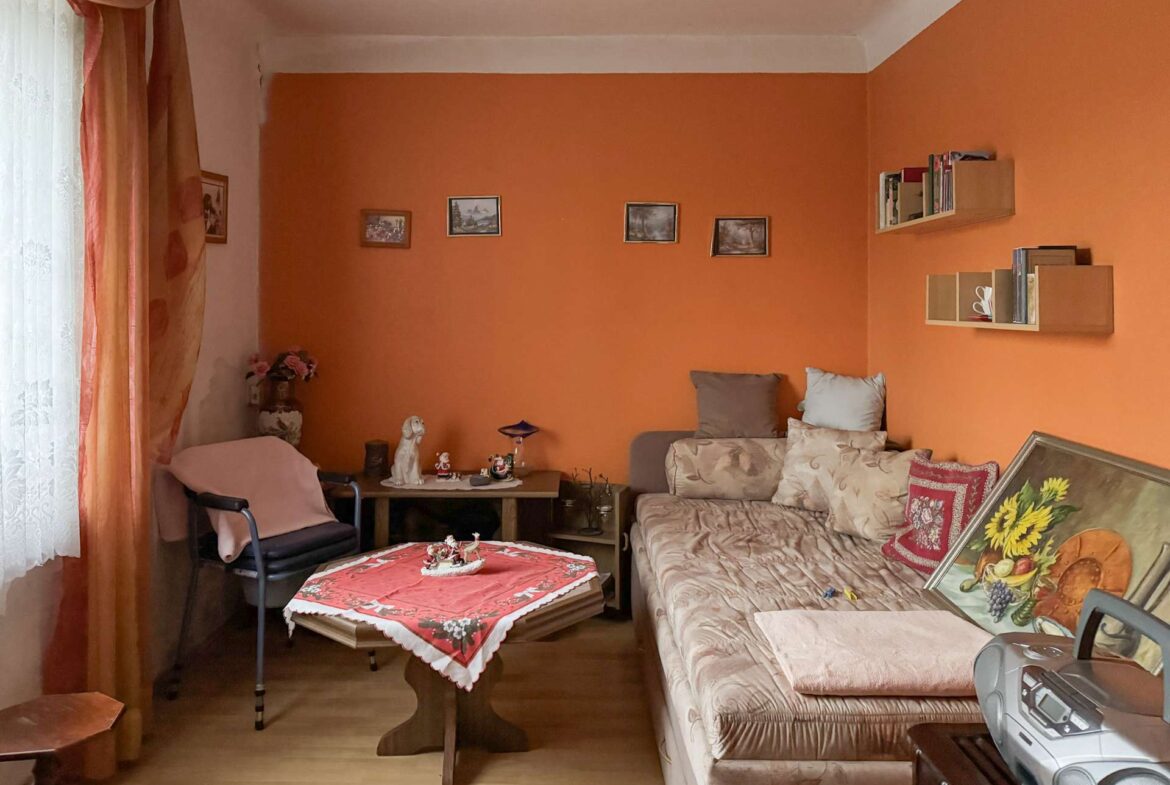 Cozy living room with an orange wall, beige sofa, patterned cushions, and a wooden coffee table covered by a red tablecloth; window with sheer white curtains on the left.