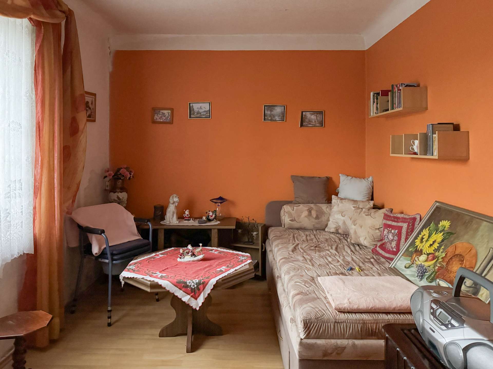 Cozy living room with an orange wall, beige sofa, patterned cushions, and a wooden coffee table covered by a red tablecloth; window with sheer white curtains on the left.