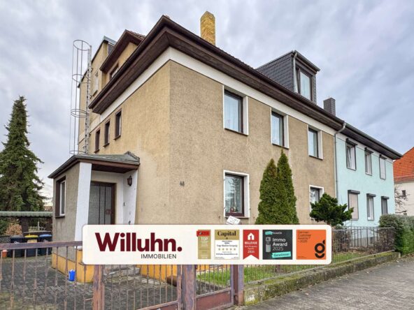 Three-story beige residential building with a large real estate sign in the foreground reading 'Willuhn Immobilien' and award stickers.