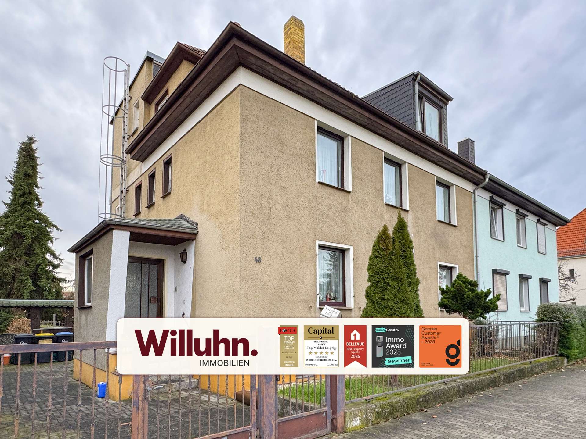 Three-story beige residential building with a large real estate sign in the foreground reading 'Willuhn Immobilien' and award stickers.