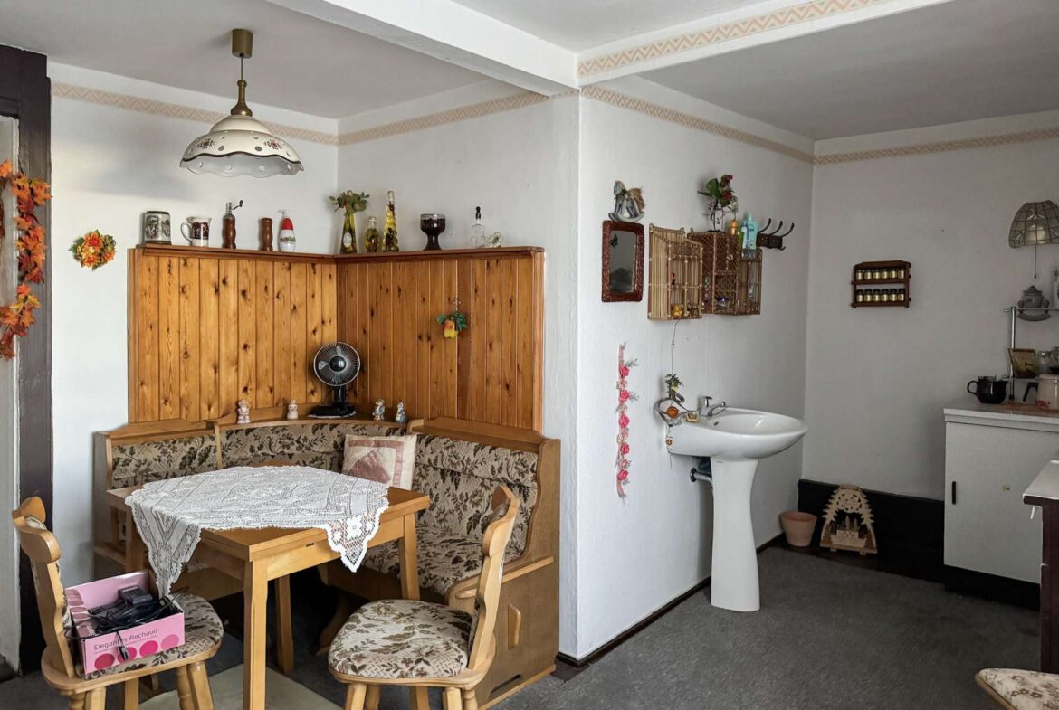 Cozy dining nook with a wooden cabinet, lace tablecloth, and floral-upholstered chairs in a vintage-style kitchen area of a home.